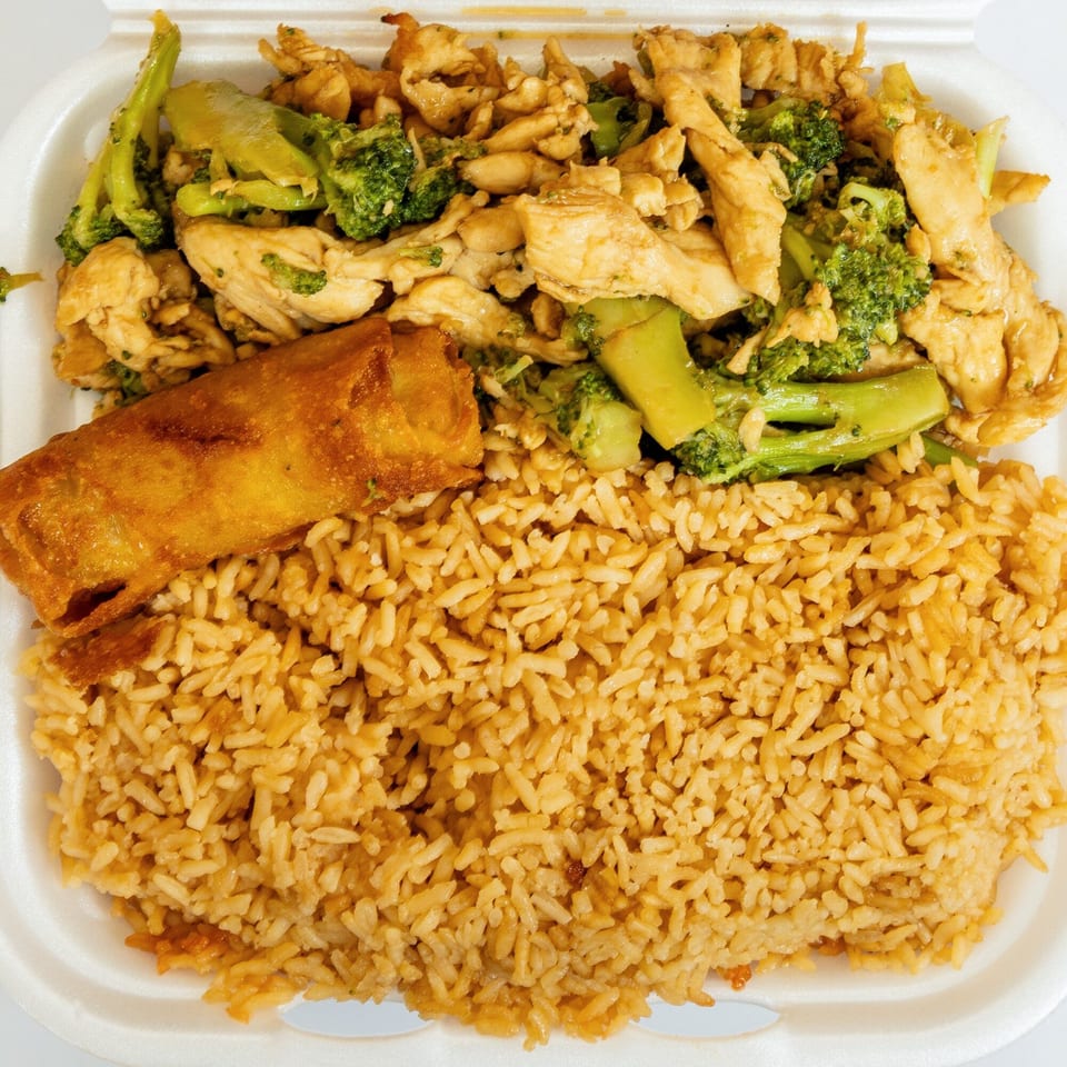 Chicken and broccoli with fried rice and an egg roll in a white takeout container.