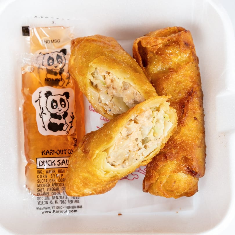 Golden-brown egg rolls, one cut open to show the filling, next to a packet of duck sauce.